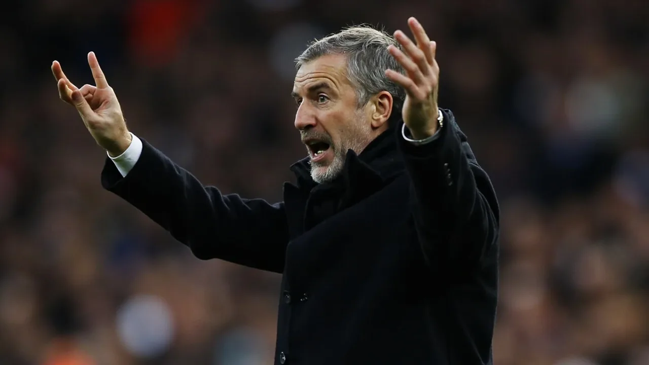 Roberto De Zerbi in a dark coat on the touchline at the Tottenham Hotspur Stadium, gesturing with one arm raised during a Premier League match in autumn daylight.