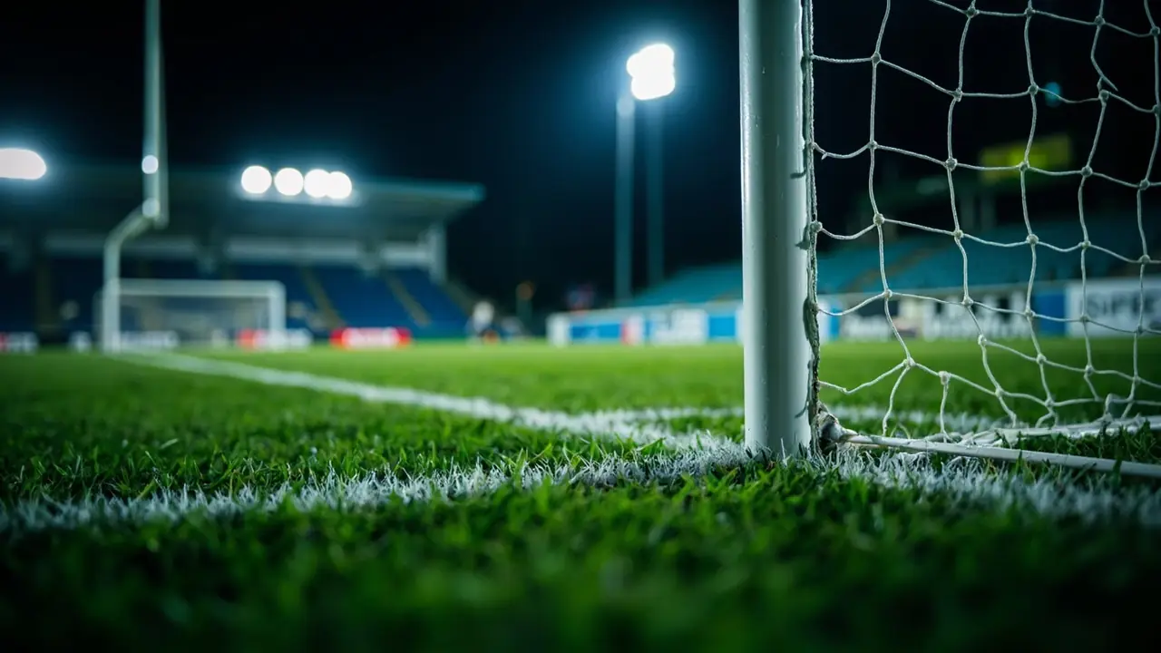 A white goalpost and net at the far post under floodlights at night, with damp grass and the six-yard line in the foreground