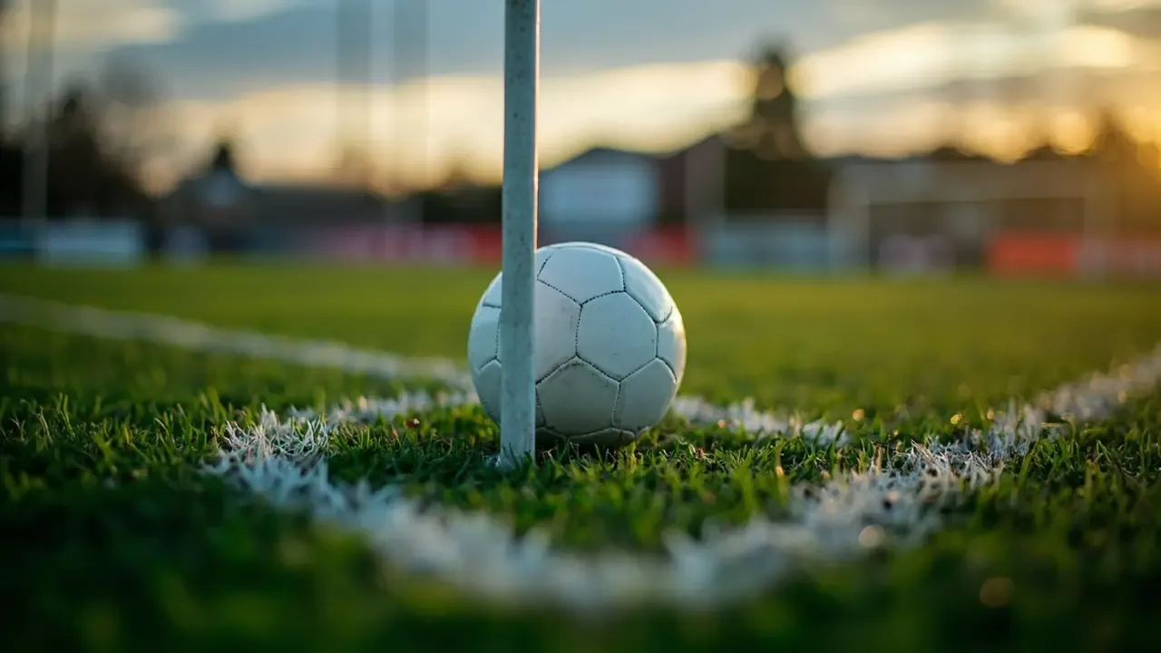 A white football resting inside the chalk corner arc beside the base of a corner flag, with the rest of the pitch blurred in evening light